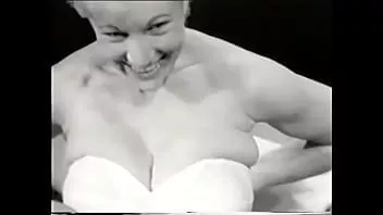 Play Sexy blonde in a white corset poses for the camera exposing her big tits to a photographer from the 60s for a men's magazine
